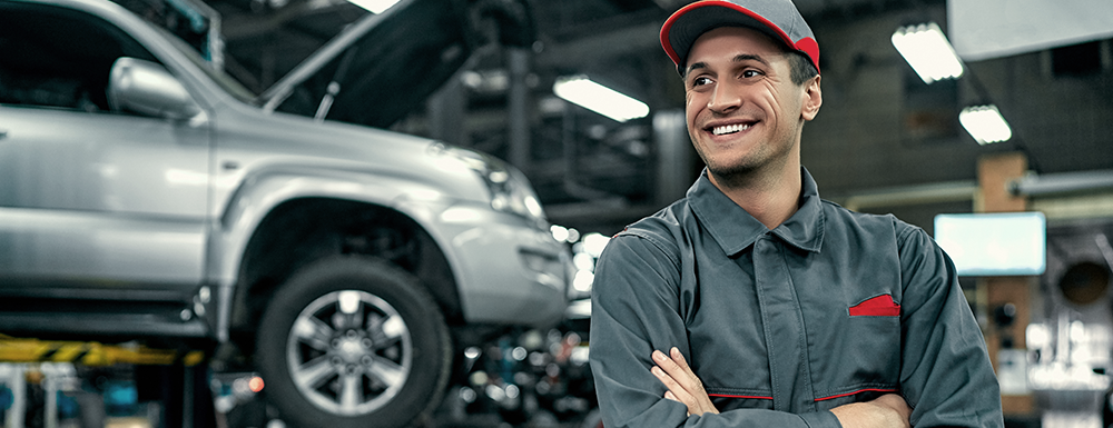 Auto service mechanic in uniform is standing on the background of car with open hood, smiling and looking away. Car repair and maintenance. Top auto franchises.