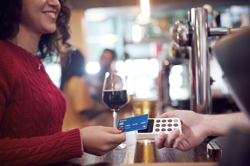 Woman at a bar scans credit card with a glass of wine by her side - Comfort items and vices, like alcohol, are often recession proof when it comes to business.