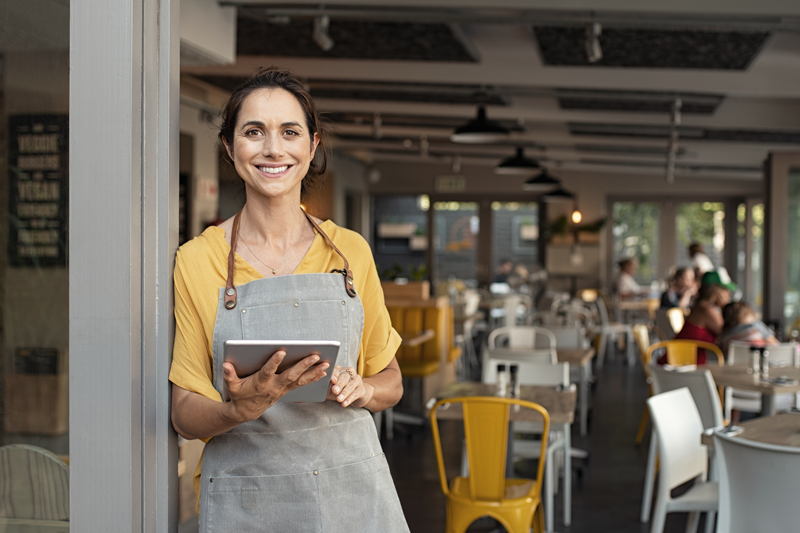 Woman wearing an apron and holding a device smiles at the camera with a restaurant scene behind her - The food and beverage industry is commonly known to be recession proof. 