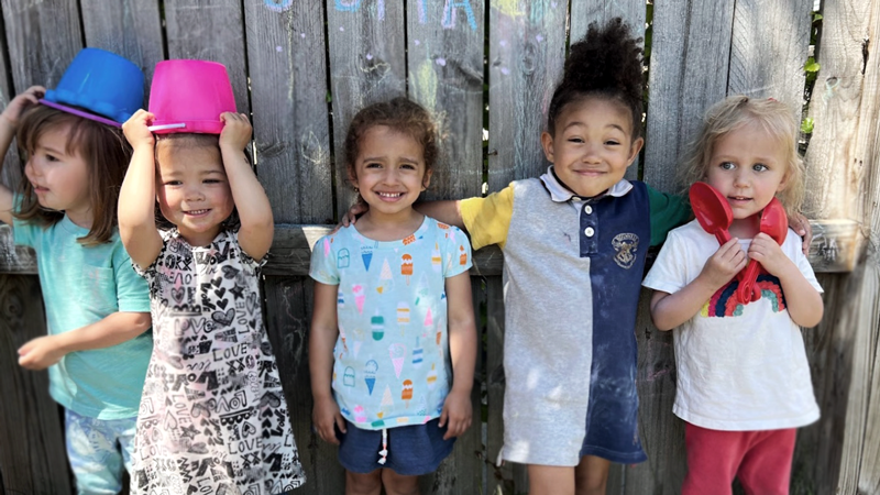 Le Village Co-Work is one of the top childcare franchises of 2022 because it offers a unique co-working and daycare option for parents. Image shows five kids playing outside of Le Village Co-Work, smiling and posing for a picture.