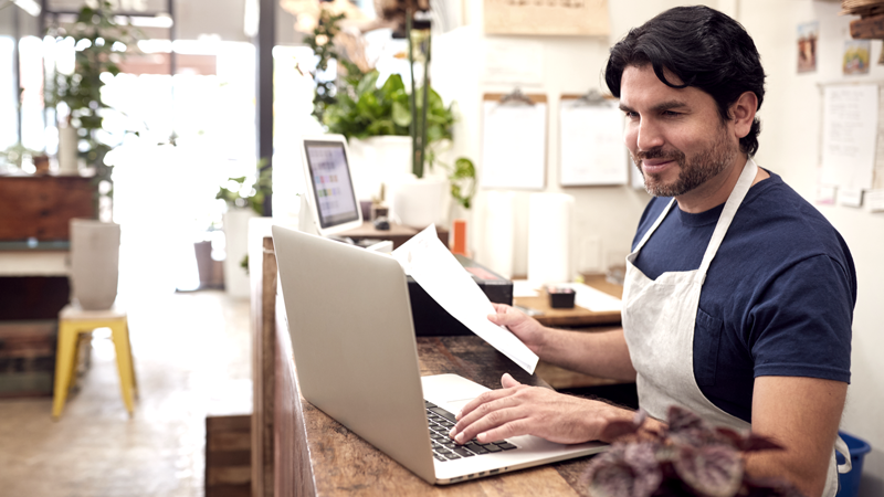 Small business owner in apron smiles while holding paperwork, implying he's doing or reviewing a small business valuation. 