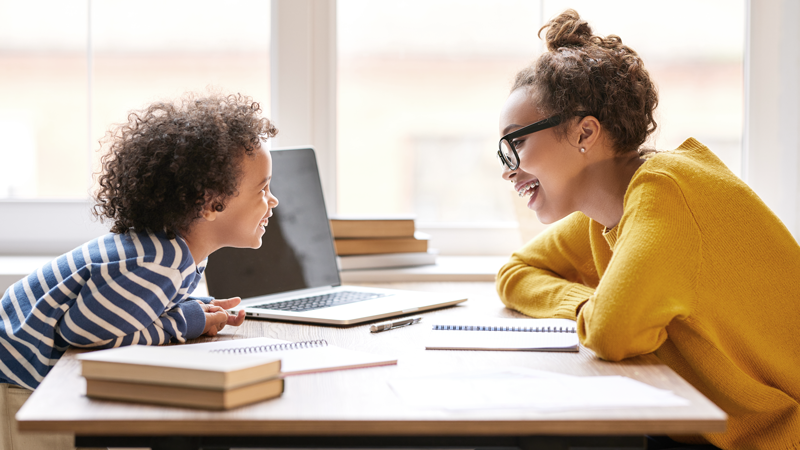 A younger child and an older child laugh while working on schoolwork together. 