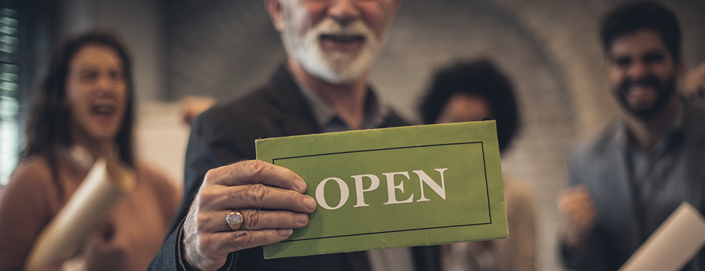 Business owner holds an "open" sign. Blog header for 10 Steps to Starting a Business.