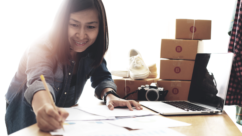 A data-driven business owner analyzing paperwork on table with pencil on a desk with a laptop and shipping boxes. 