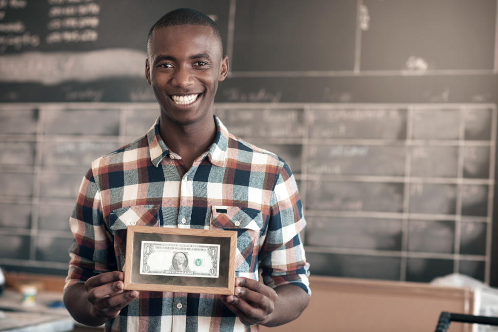Business owner holding his first dollar in a frame, smiling. (Small business vs Startup: What's the difference between startup and small business - Blog). 