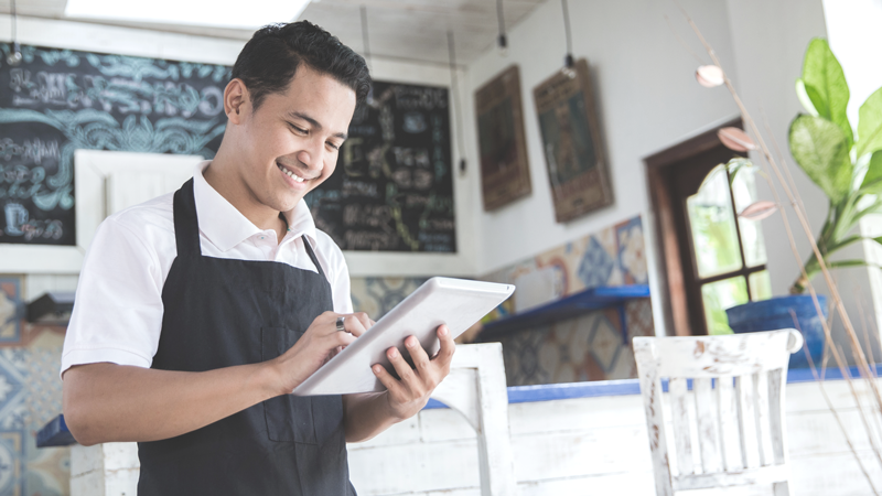 Smiling business owner in a cafe wearing an apron looks at tablet. 