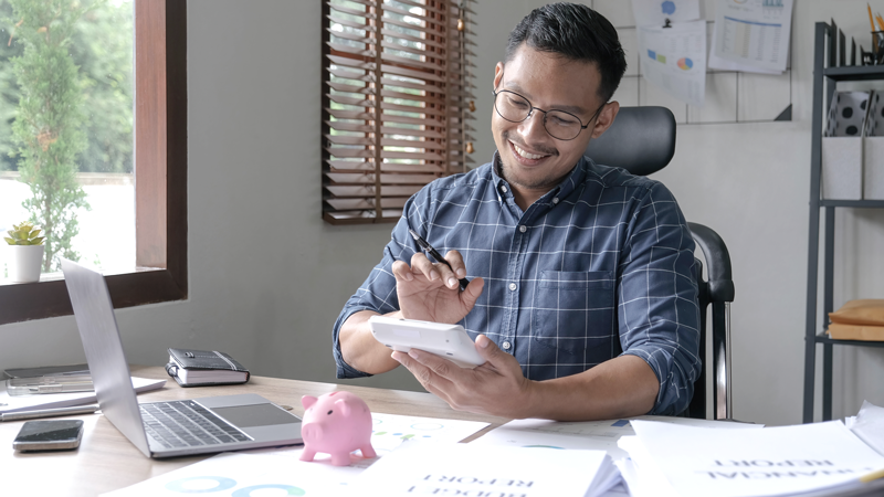 Man doing finances while smiling in his office at his desk, topped with a piggy bank, laptop, and various paperwork. (Top Resources for Getting Started with ROBS Blog.)