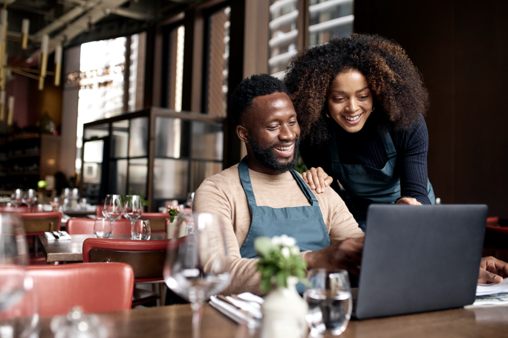 Black business owners of a restaurant looking at a laptop screen together and smiling. 