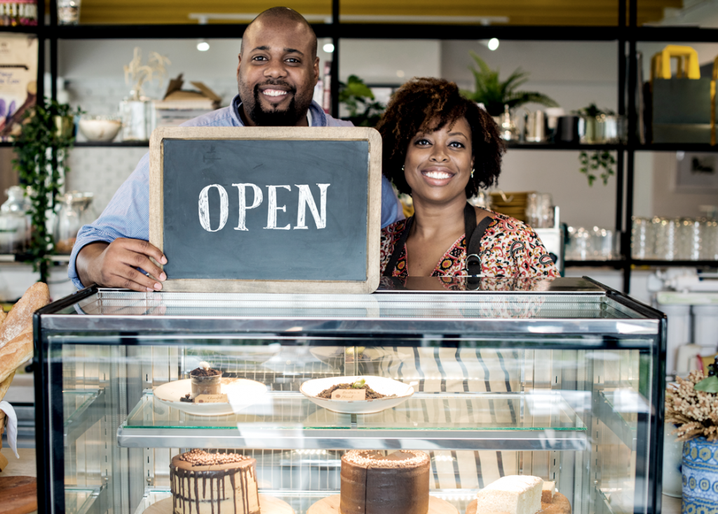 An Black entrepreneur couple holding an open sign on top of a case filled with cakes and other bakery-themed items.  (Top Black Entrepreneur Grants and Resources Blog.) 