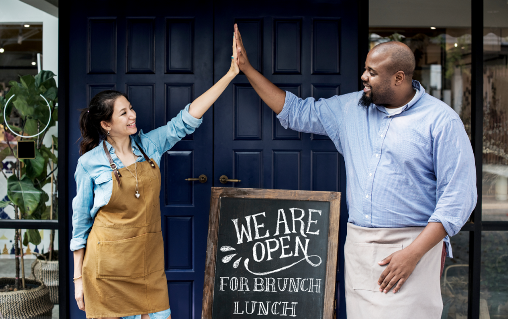 Two business owners high-fiving outside a restaurant storefront wearing aprons. (5 Black Business Owners Making Waves Blog). 