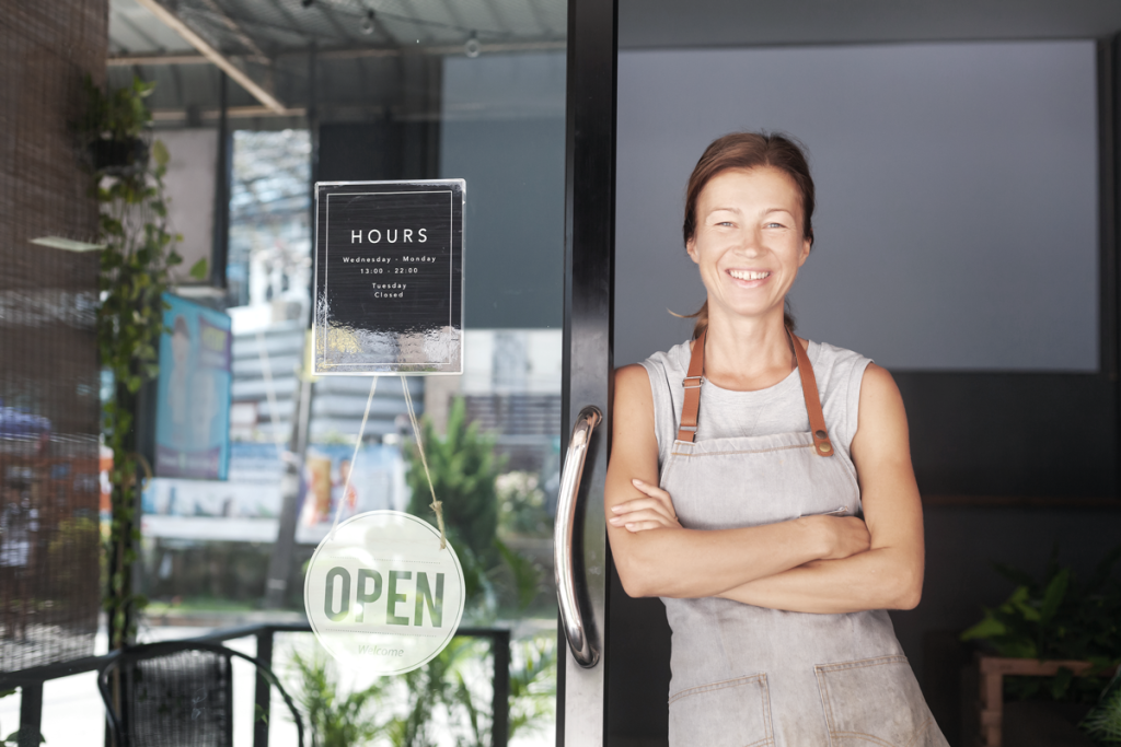 Woman business owner standing proudly outside of storefront smiling next to a hanging "OPEN" sign. (Top Resources for Getting Started with ROBS Blog.)