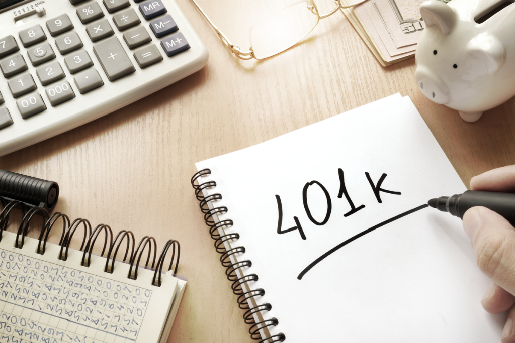 A notebook on a desk reads "401k" next to a piggy bank, calculator, money, and paperwork. (10 Tax Benefits of C Corporations - Guidant Blog.)