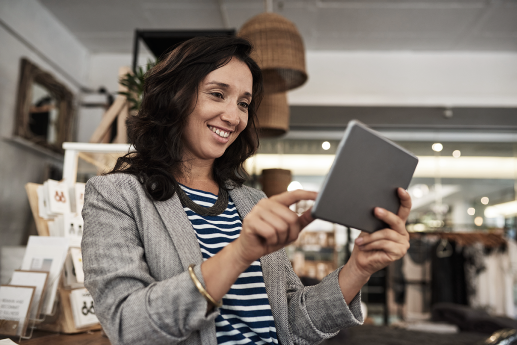 Woman business owner holding a tablet and smiling inside a clothing store. (Business Grants for Women Blog - Guidant). 
