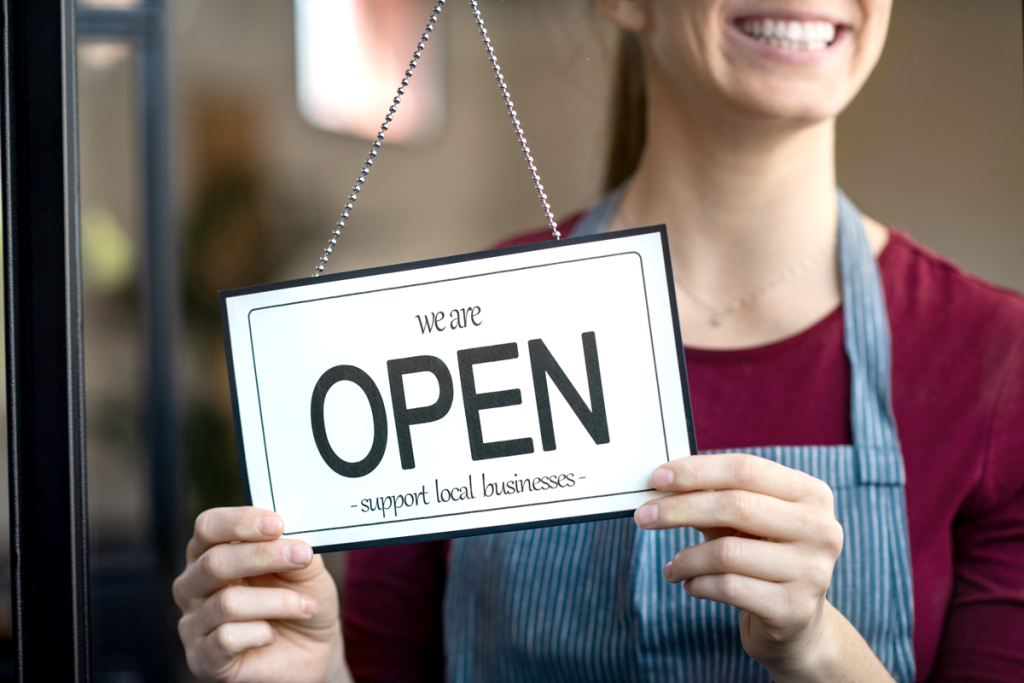 Business owner smiling and holding open a sign that says "We are open, support local businesses" (Ways to Celebrate Small Business Week - Guidant Blog)