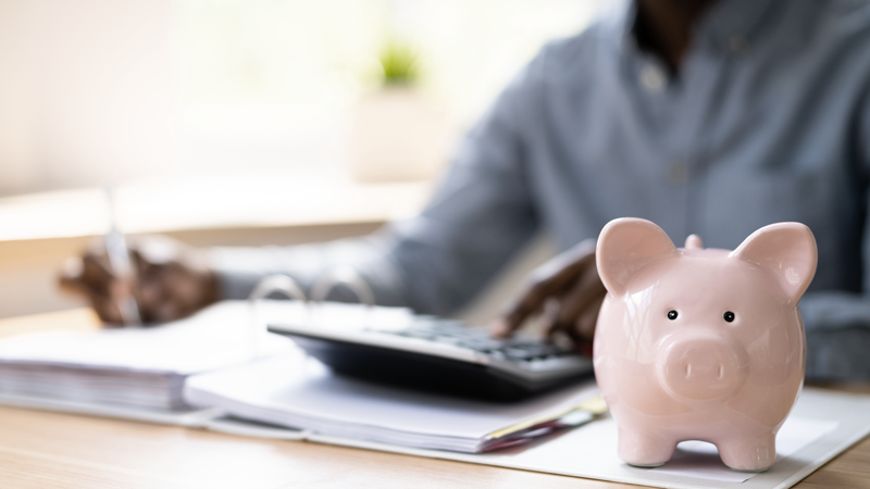 Man in the background at his desk doing finances with a calculator and a piggy bank next to him. (Blog: Small Business Week, Big Dreams: How to Find and Secure Business Financing)