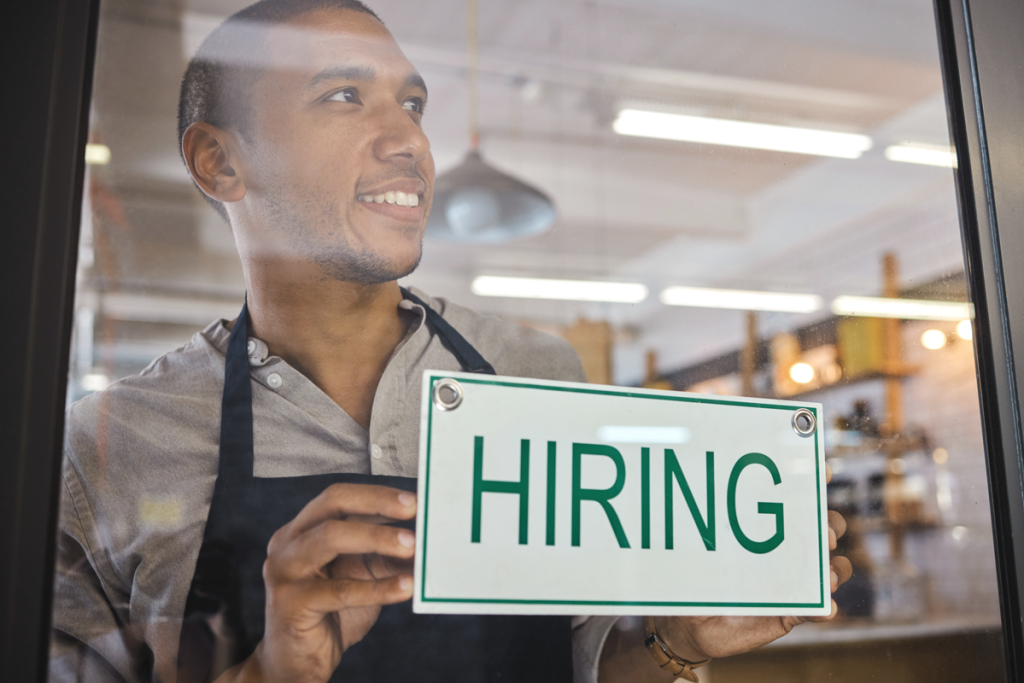 Man holding a hiring sign from inside his store. (Hiring for Small Business 101: When and How to Find the Right Talent - Guidant Blog). 