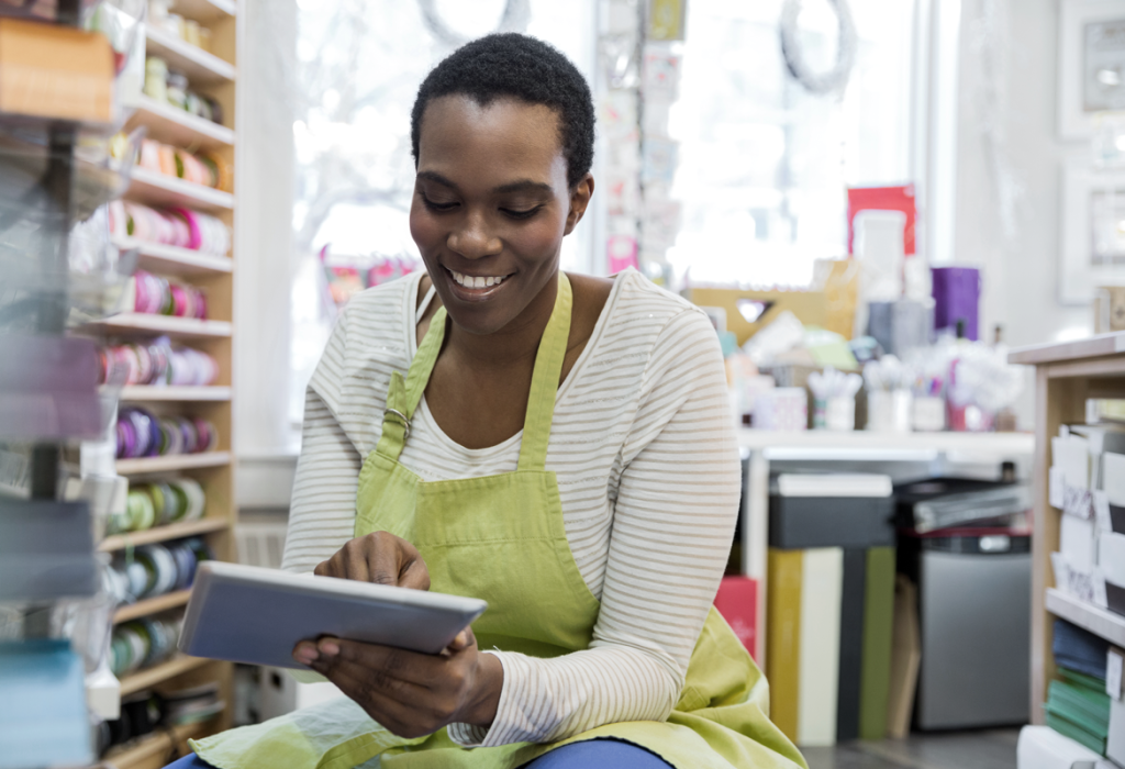 Woman business owner in an apron inside her shop, planning on her tablet.  (Tips on How to Run Your Small Business on Summer Vacation - Guidant Blog.)