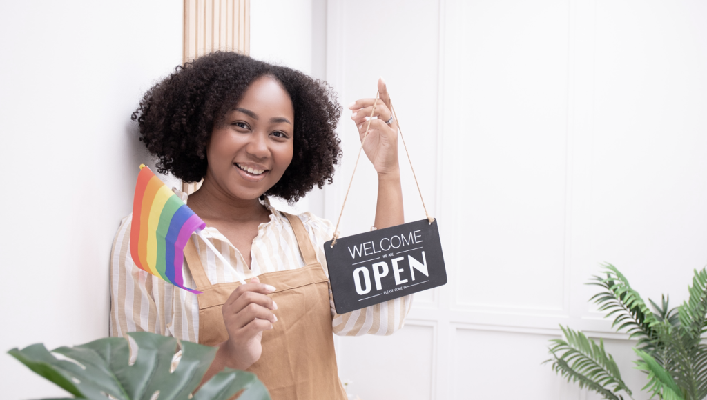Small business owner Black woman holding a "Welcome, we're open" sign and a rainbow pride flag. (5 Ways Small Businesses Can Support Pride Month - Guidant Blog.)