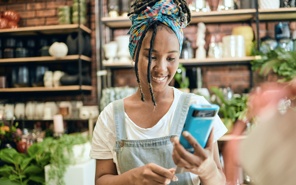 Black woman small business owner smiling and looking at her mobile phone while at work. (Turn Up the Heat with these Top 7 Summer Sale Promotion Ideas for Small Businesses - Guidant Blog.)
