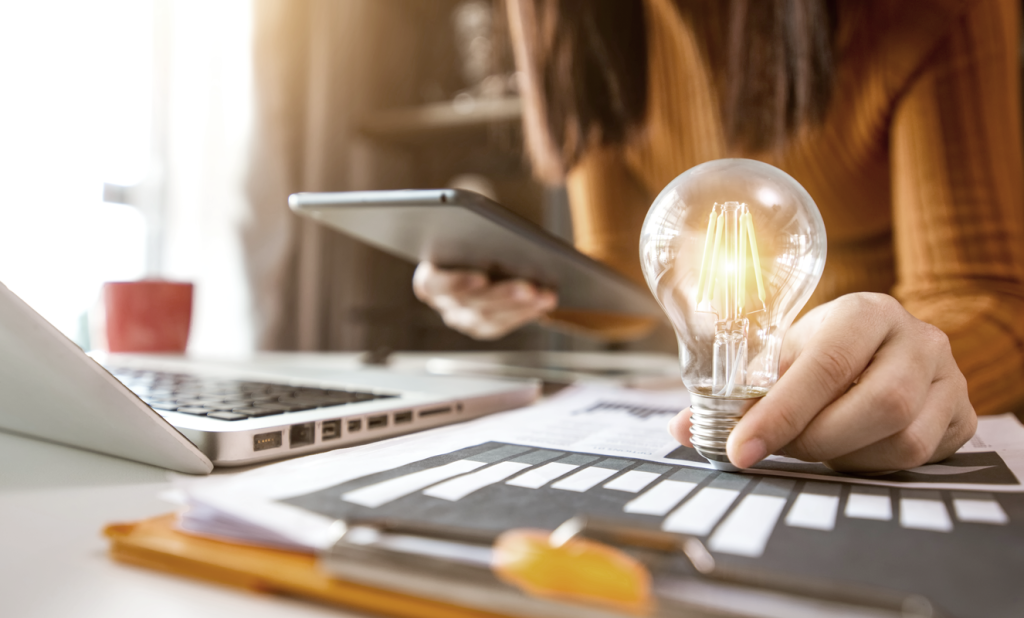 Woman at her workspace holding a lit light bulb while working on a laptop and tablet. (Transitioning from Office to Entrepreneurship: How to Start a Business with these Top 3 Startup Strategies - Guidant Blog.)