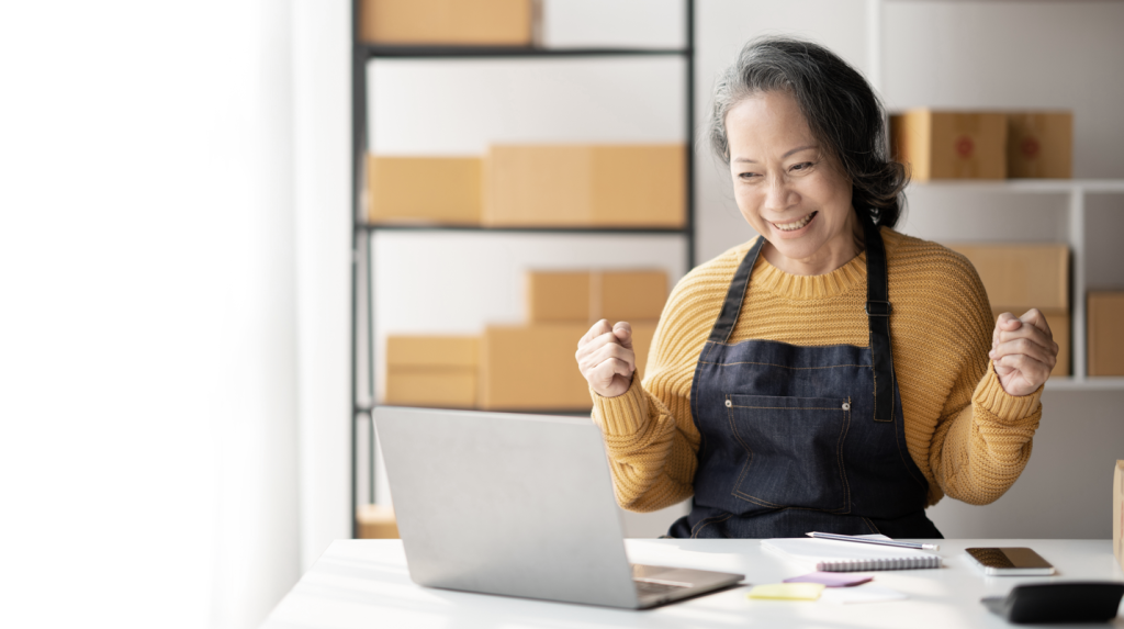 Asian woman business owner wearing an apron looking excited at desk (7 Best Options for Small Business Funding - Guidant Blog). 