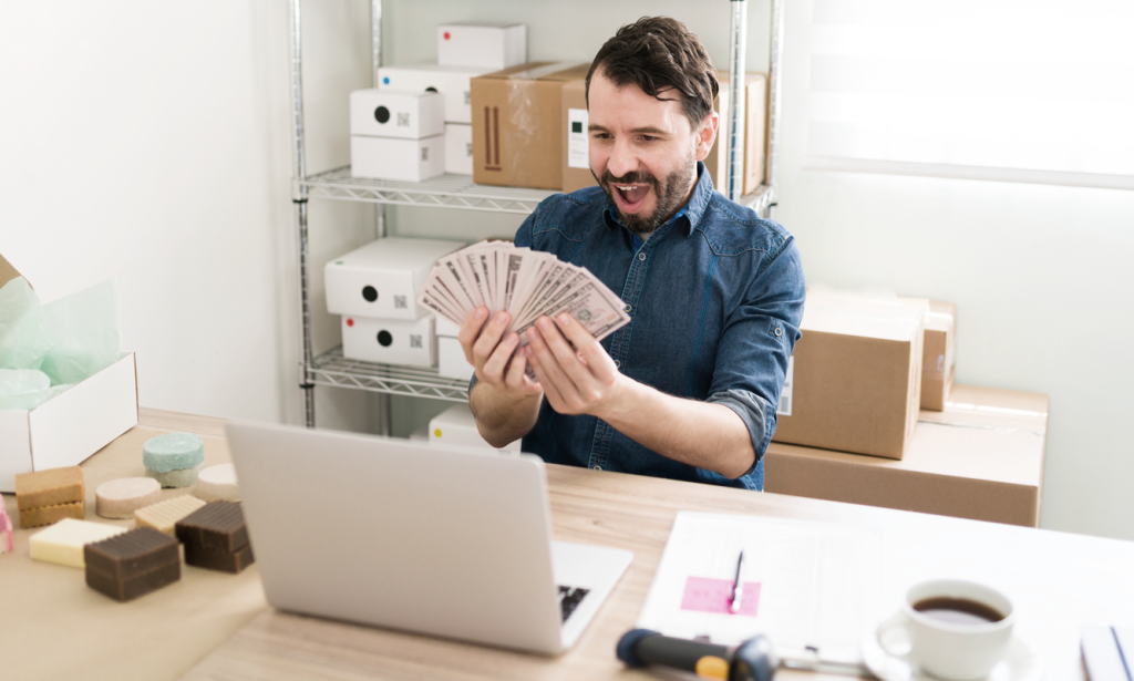 Business owner at desk looking excited white holding cash. (7 Best Options for Small Business Funding - Guidant Blog). 