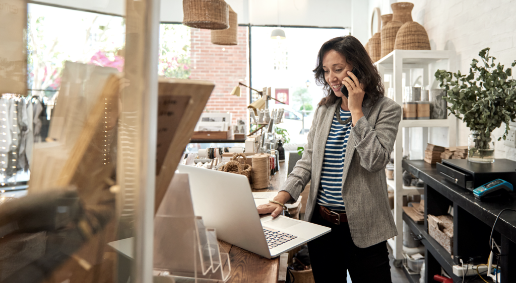 Woman business owner on the phone while looking at a laptop while in a retail store. (Transitioning from Office to Entrepreneurship: How to Start a Business with these Top 3 Startup Strategies - Guidant Blog.)