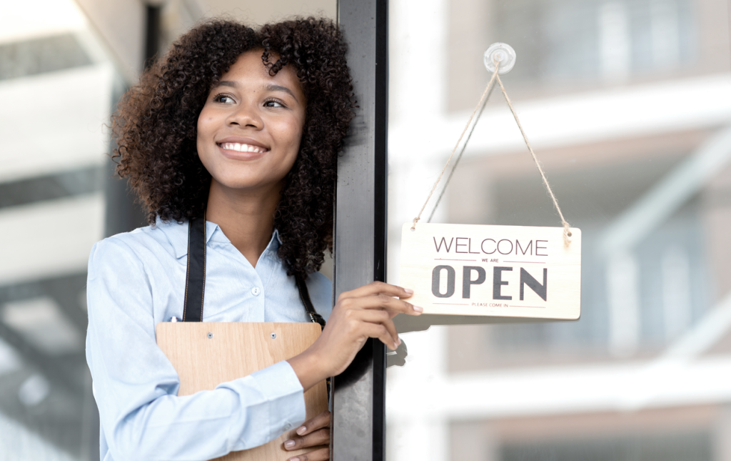 Business owner standing outside store location holding a "Welcome, we're open" sign. (Transitioning from Office to Entrepreneurship: How to Start a Business with these Top 3 Startup Strategies - Guidant Blog.)