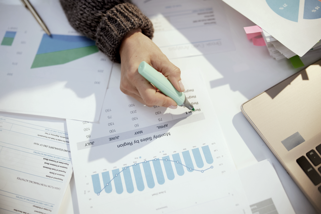 A desk filled with paperwork of sales and revenue data for a business, and a hand highlighting figures. (Inflation-Proofing Your Small Business: Insights and Tactics from Successful US Entrepreneurs - Guidant Blog).