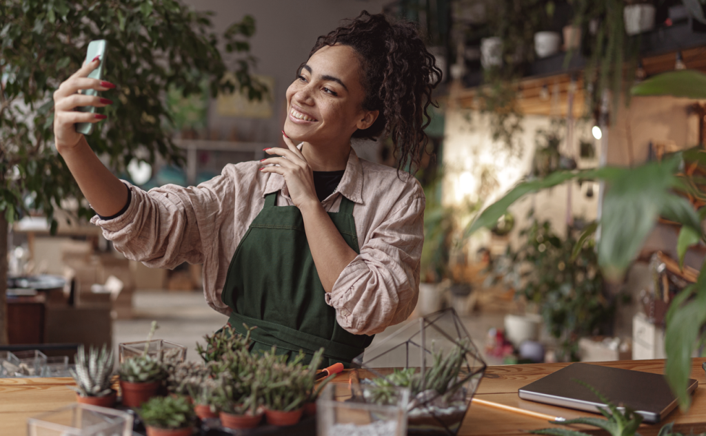 Woman business owner holding a phone and taking a selfie while working in a location filled with plants. (Boost Your Small Business with the Power of Hockey Stick Growth in 7 Steps - Guidant).