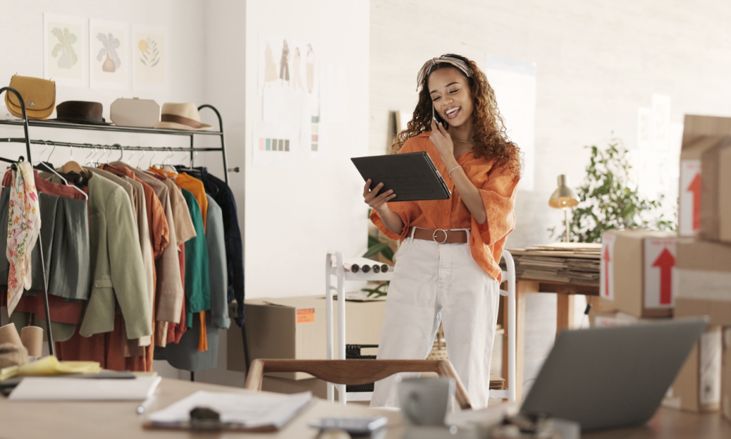 Woman happily talking on the phone while looking at a tablet in a fashion and clothing studio space.  (Top 5 Surprising Benefits of Offshoring for Small Businesses - Guidant)