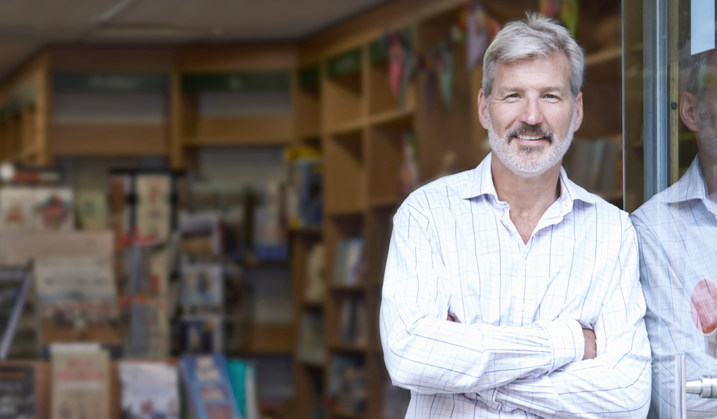 Older male business owner smiling confidently outside business store front. (What is ROBS? How 401k Business Financing Works - Guidant Blog).