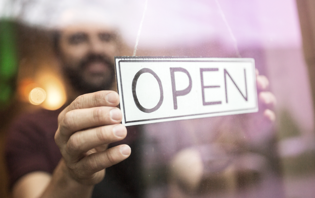 Male business owner holding an open sign in the door front of his business. (Top Resources and Grants for Hispanic Entrepreneurs - Guidant Financial Blog).
