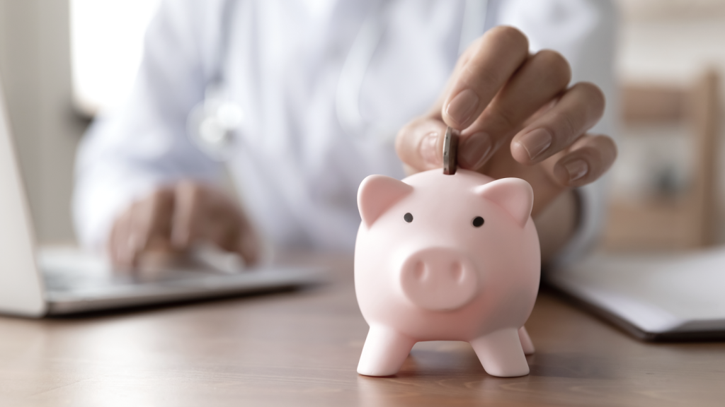 Person at desk in background putting a coin into a piggy bank while on laptop. (Demystifying Top Heavy 401k Plans: What You Need to Know - Guidant Blog.)