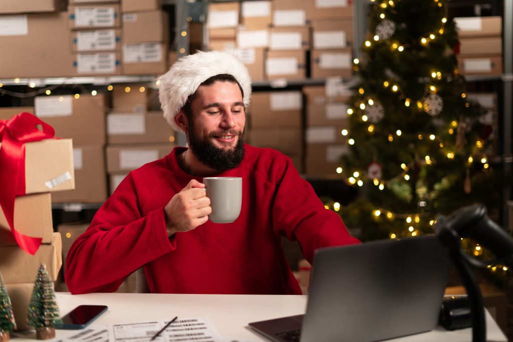 Manager checking online orders and inventory on a laptop drinking coffee or tea while sitting at a desk in warehouse with shelves of stock on background. (How To: Small Business Marketing for the Holidays - Guidant). 