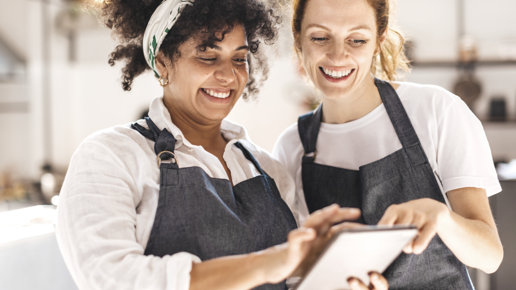 Business owner and employee in aprons smiling and using a tablet. (Top 5 Must-Have New Year's Resolutions for Your Small Business - Guidant Blog.)