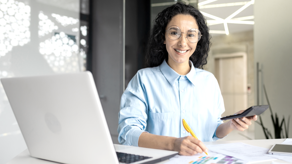 Happy woman smiling at desk holding a calculator and pencil. (Complete Guide to Payroll for Small Businesses - Guidant Financial.)