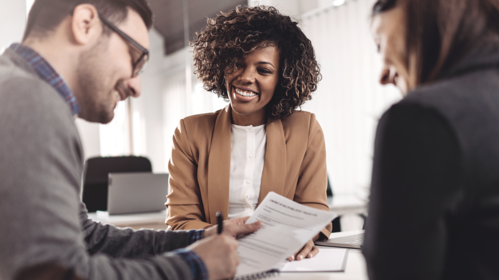 Three people smiling while signing paperwork in an office setting. (Buy a Small Business with Seller Financing - Guidant Blog)