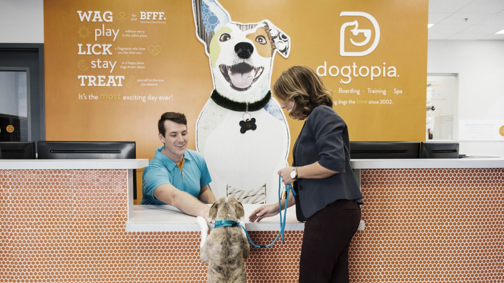 Woman and dog at the front desk of a Dogtopia store wiith a smiling employee. (Hottest Pet Care Franchises - Guidant Financial Blog).