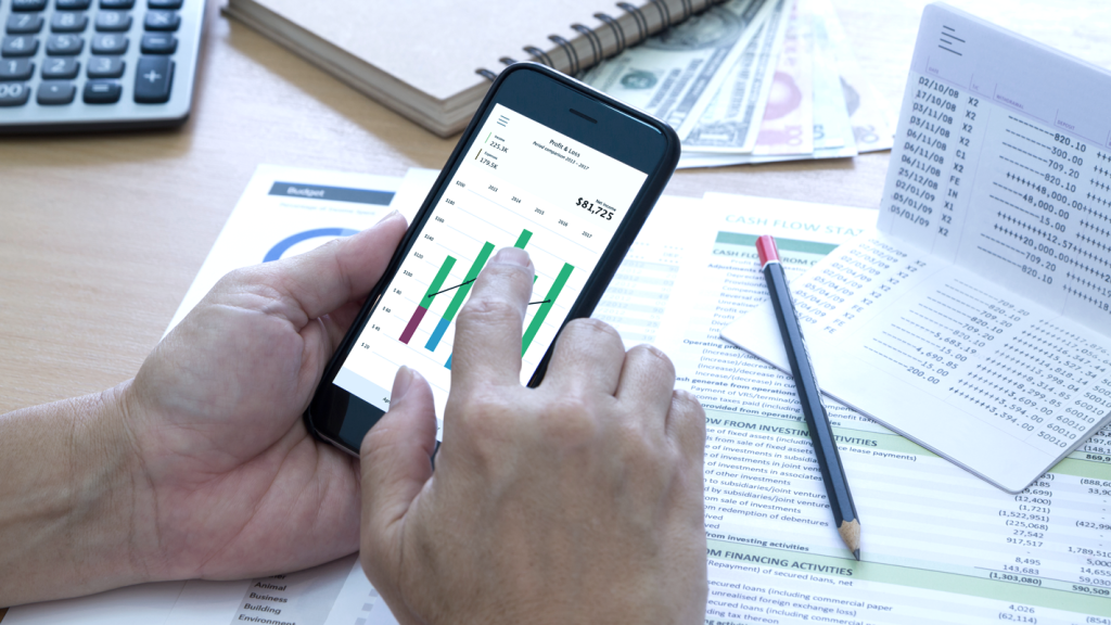 Hands using a mobile phone pointing at a graph on desk with financial paperwork. (8 Vital Cash Flow Management Tips for Small Business Owners - Guidant Blog).