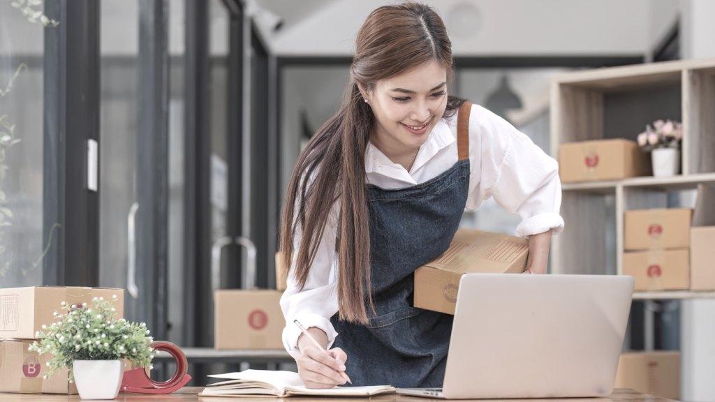 Woman business owner managing inventory, holding a box and writing in a log in an office space. (8 Vital Cash Flow Management Tips for Small Business Owners - Guidant Blog).