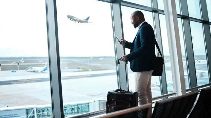 Man on his phone with a suitcase next to a window, looking out a sky view with a plane flying.  (Top Tourism Industry Trends and Top Travel Industry Trends - Guidant Financial.)